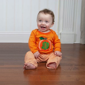 May include: A baby boy wearing an orange long-sleeve shirt with a pumpkin applique and the monogram "JDD" on it. He is also wearing orange and white checkered pants.