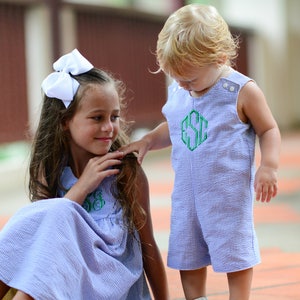 K&ouml;nnte beinhalten: Zwei Kinder tragen passende blau-wei&szlig; gestreifte Outfits mit gr&uuml;nen gestickten Monogrammen. Das M&auml;dchen tr&auml;gt eine wei&szlig;e Schleife im Haar und schaut nach rechts. Der Junge schaut das M&auml;dchen an und hat blonde Haare.