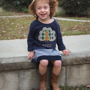May include: A young girl wearing a navy blue long-sleeved shirt with a green and white plaid turkey design and the name "Annie" embroidered on the front. She is wearing a blue and white gingham skirt and brown cowboy boots. She is sitting on a concrete wall and smiling.