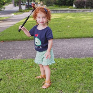May include: A young girl with curly red hair wearing a blue and green golf-themed shirt and green and white checkered shorts holds a golf club over her head.