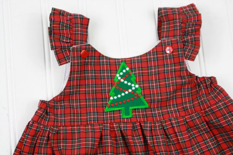 K&ouml;nnte beinhalten: Rotes und gr&uuml;nes Karo-Overallkleid mit einem gr&uuml;nen Filz-Weihnachtsbaum-Applikation. Der Baum hat wei&szlig;e und rote Perlen und einen roten Stern oben drauf.