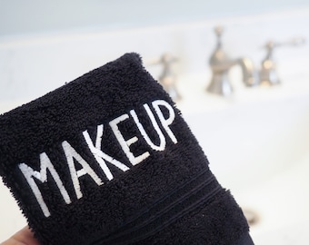 May include: A black face cloth with the word "MAKEUP" embroidered in white. The cloth is held in front of a blurred white sink and taps.