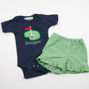 May include: A navy blue baby onesie with a green and white golf course design and the name "Reagan" embroidered on the front. The onesie is paired with green and white gingham shorts with a ruffled hem.