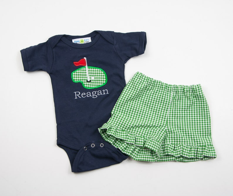 May include: A navy blue baby onesie with a green and white golf course design and the name "Reagan" embroidered on the front. The onesie is paired with green and white gingham shorts with a ruffled hem.