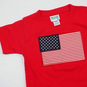 May include: Red short-sleeved t-shirt with an embroidered American flag on the front. The flag is in the top right corner of the shirt. The shirt has a crew neck and is made of 100% cotton. The tag on the shirt says "Soda City Sewing" and "3/4".