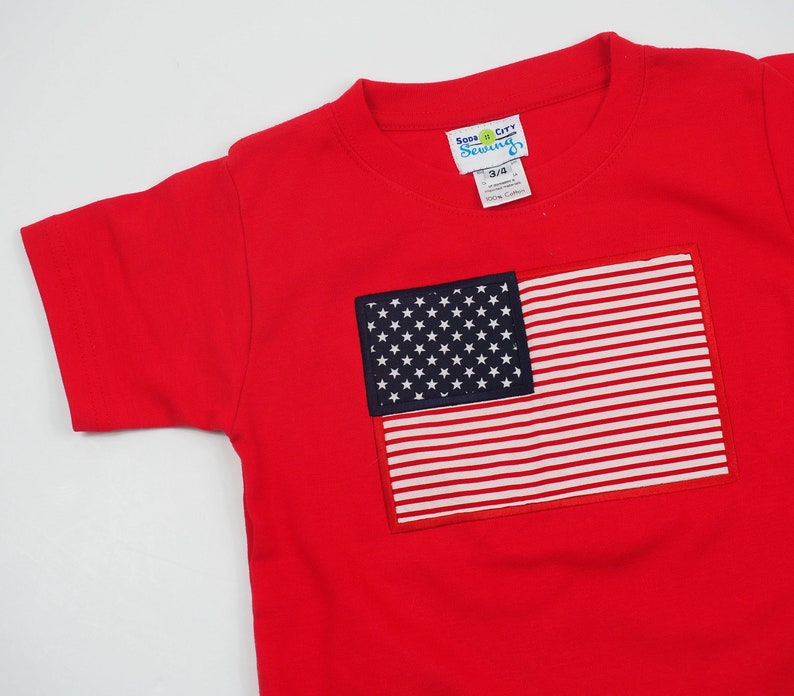 May include: Red short-sleeved t-shirt with an embroidered American flag on the front. The flag is in the top right corner of the shirt. The shirt has a crew neck and is made of 100% cotton. The tag on the shirt says "Soda City Sewing" and "3/4".