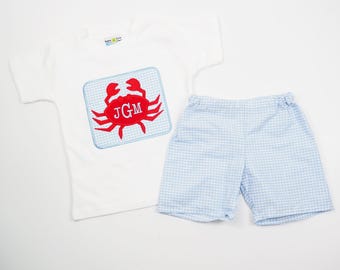 Puede incluir: Conjunto de camiseta blanca y pantalones cortos de cuadros vichy azul claro. La camiseta presenta un aplique de cangrejo rojo con las iniciales "JGM" sobre un fondo de cuadros azules y blancos. Los pantalones cortos son de cuadros vichy azul claro y blanco.