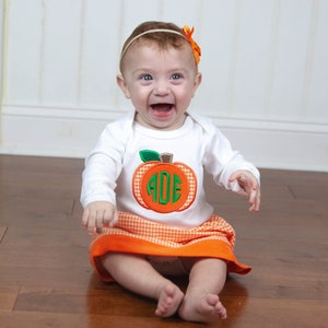 Op de afbeelding: Een babymeisje draagt een wit shirt met lange mouwen met een oranje en groene pompoenapplicatie. De pompoen heeft de letters "ADE" in groen erop. De baby draagt ook een oranje en witte geruite rok. De baby zit op een houten vloer en glimlacht.