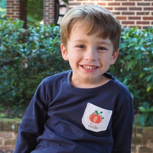 May include: A young boy wearing a navy blue long-sleeved shirt with a white patch pocket featuring an embroidered pumpkin and the name "Jude".