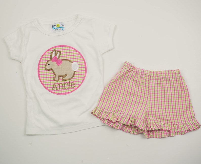 Op de afbeelding: Wit T-shirt met korte mouwen met een roze en groene ginghamcirkel met een geborduurde bruine konijn met een roze strik. De cirkel heeft de naam "Annie" onder het konijn. Het shirt wordt gecombineerd met roze en groene gingham shorts met een geruite zoom.