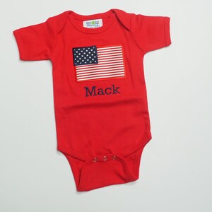 May include: Red short-sleeved baby onesie with a blue and white American flag and the name "Mack" embroidered on the front.