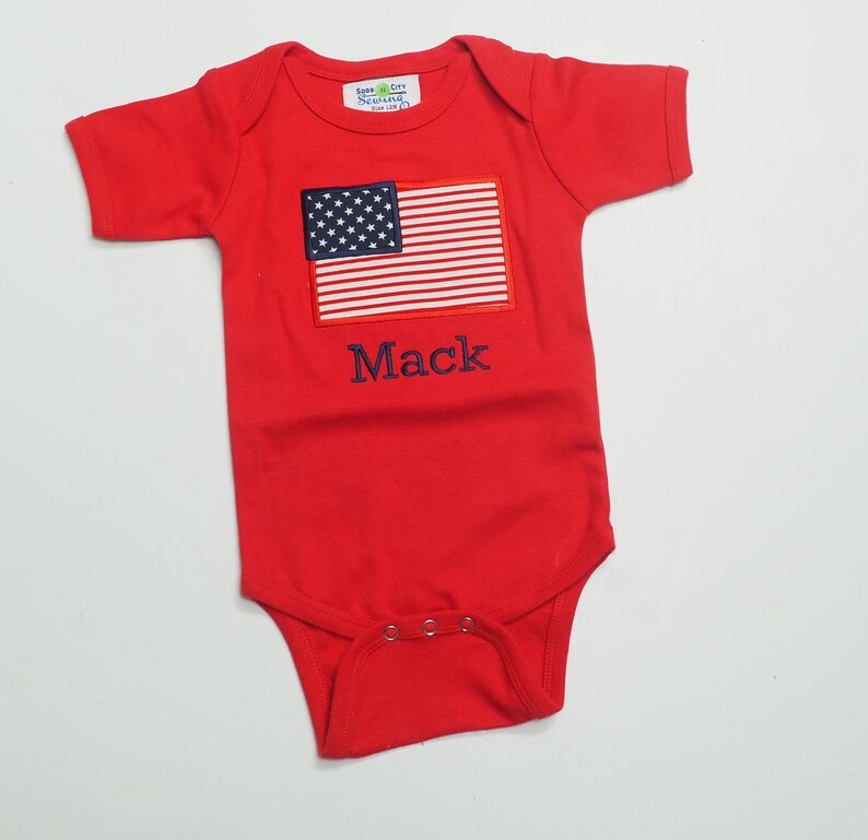 May include: Red short-sleeved baby onesie with a blue and white American flag and the name "Mack" embroidered on the front.