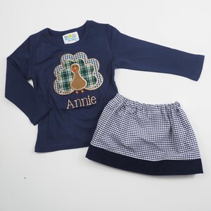 May include: A navy blue long-sleeved shirt with a plaid turkey applique and the name "Annie" embroidered on the front. The shirt is paired with a white and navy blue gingham skirt with a navy blue waistband.