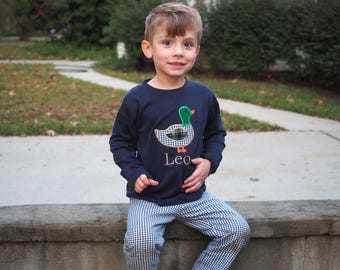 Puede incluir: Un niño pequeño con una camisa de manga larga azul marino con un aplique de pato verde, blanco y naranja. La camisa tiene el nombre "Leo" bordado en la parte delantera. Lleva pantalones a cuadros azules y blancos.