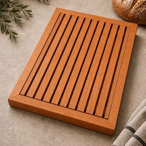 May include: A rectangular wooden bread cutting board featuring a slatted surface design with a solid frame border. The board is constructed from light brown wood with a visible grain pattern, designed with open gaps between the slats for crumbs.
