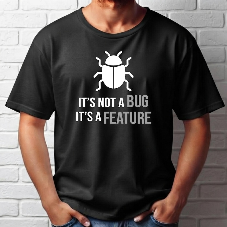 Może przedstawiać: Czarny t-shirt z kr&oacute;tkim rękawem z białą grafiką przedstawiającą ikonę chrząszcza nad napisem IT'S NOT A BUG IT'S A FEATURE wydrukowanym białymi wielkimi literami. Koszulka ma okrągły dekolt i standardowy kr&oacute;j.