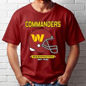 May include: A burgundy short-sleeve t-shirt featuring a graphic of a football helmet with a yellow W logo. The text COMMANDERS is printed above the helmet, and WASHINGTON EST 1932 is printed below the helmet in yellow and white lettering.