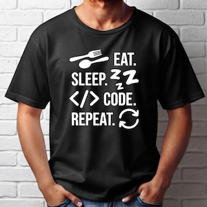May include: A black short-sleeved t-shirt featuring white graphic text and icons. The design includes a fork and spoon icon next to the word EAT, the word SLEEP with Z symbols, code brackets next to the word CODE, and the word REPEAT with a cycle icon.