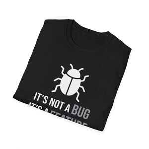 Może przedstawiać: Czarny t-shirt z białą grafiką przedstawiającą ikonę chrząszcza nad białym napisem IT'S NOT A BUG IT'S A FEATURE. Koszulka ma okrągły dekolt i kr&oacute;tkie rękawy, zaprezentowana w złożonej, płaskiej orientacji.
