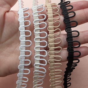 May include: Four strips of loop trim in white, off-white, beige, and black. Each strip features a woven flat base with a continuous row of uniform, rounded fabric loops extending from one side, designed for button closures or decorative edging.