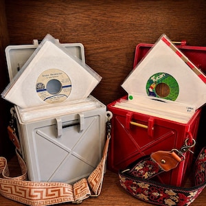 May include: Two square storage cases, one light gray and one red, each holding vinyl records in clear sleeves. The gray case has a tan patterned strap, while the red case features a black and red floral strap. Both cases have a cross-patterned exterior.