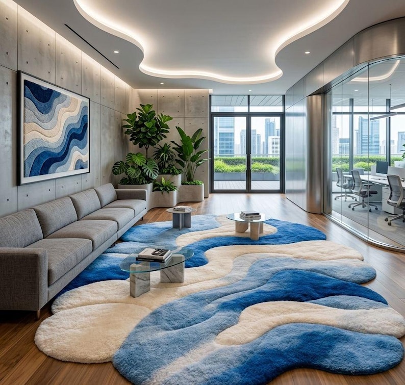 May include: A large, irregular, wavy-shaped area rug featuring a gradient of blue, light blue, and cream colors. The rug is placed on a wooden floor in a modern office space with a gray sofa, glass coffee tables, and potted green plants.