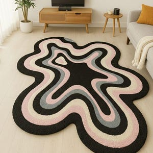 May include: An irregular, blob-shaped area rug featuring concentric, wavy bands of color. The design consists of alternating layers of black, soft pink, light gray, and cream, creating a fluid, organic pattern across the textured surface.