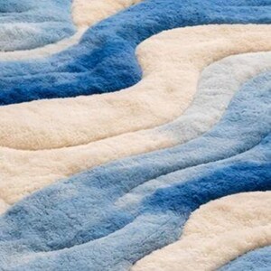 May include: A plush area rug featuring a wavy, abstract pattern of alternating bands in shades of deep blue, light sky blue, and creamy off-white. The surface has a thick, soft, and textured pile that creates a dimensional, rippled appearance.