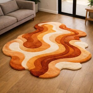 May include: An irregularly shaped area rug featuring a wavy, fluid pattern in shades of cream, light orange, and deep burnt orange. The rug has a thick, textured pile and organic, flowing curves that create a layered, multi-colored effect.