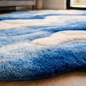 May include: A plush area rug featuring a wavy, abstract pattern in shades of deep blue, light blue, and cream. The rug has a thick, tufted texture with high-pile sections that create a dimensional, rippled effect across the entire surface area.