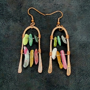 May include: A pair of copper earrings featuring a hammered arch frame with two horizontal wires. Each earring is adorned with small, irregular, multi-colored gemstone chips in shades of green, yellow, pink, white, and black, suspended from hooks.