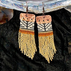 May include: A pair of dangle earrings featuring hammered copper rectangular tops with wire hooks. The design includes rows of small seed beads in white, black, and orange, transitioning into long, flowing fringe strands of metallic gold beads.