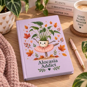 Könnte beinhalten: Ein hellviolettes Notizbuch mit festem Einband, das eine Illustration einer Alocasia-Pflanze im Topf als Ballerina in einem rosa Tutu zeigt, umgeben von orangefarbenen Herbstblättern und kleinen Herzen, mit dem Text Alocasia Addict darunter.