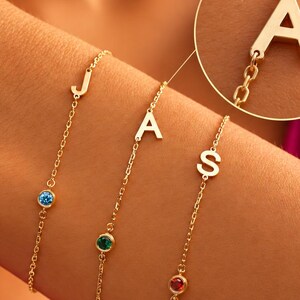Könnte beinhalten: Drei goldfarbene Kettenarmbänder, jedes mit einem Buchstabenanhänger und einem kleinen runden Edelstein. Die Buchstaben J, A und S sind zu sehen, mit blauen, grünen und roten Steinen. Ein kreisförmiger Ausschnitt zeigt eine Nahaufnahme des Buchstabens A.