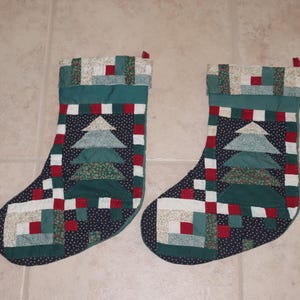 May include: Two quilted Christmas stockings featuring a patchwork design with a central pine tree motif. The stockings are constructed from dark navy, forest green, red, and cream fabric squares, with a decorative cuff at the top of each piece.