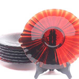 May include: A stack of round, deep red glass plates with a scalloped edge and a sunburst pattern radiating from the center. One plate is displayed upright on a black plastic stand, while the others are stacked horizontally behind it.