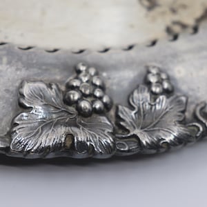 May include: Close-up of a silver-toned plate with grape and leaf embellishments. The plate's edge features a decorative border. The grapes and leaves are raised, adding texture to the design. The plate has a vintage look.