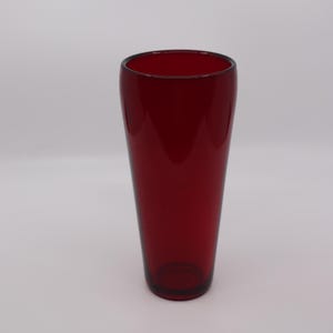 May include: A tall, ruby red glass vase with a slightly flared shape. The vase has a smooth, glossy surface and a simple, elegant design. Perfect for displaying flowers or as a decorative accent piece. The vase is approximately 8 inches tall.