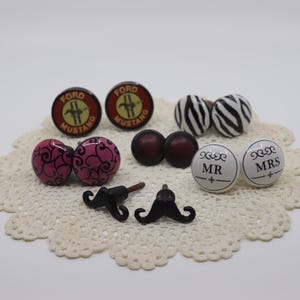 May include: A collection of decorative cabinet knobs and drawer pulls. The knobs feature various designs, including Ford Mustang logos, pink swirls, zebra stripes, solid burgundy, and "Mr" and "Mrs" text. Also included are two black mustache-shaped pulls.