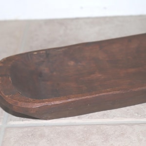 May include: A long, narrow, dark brown wooden dough bowl with a smooth, rounded interior and a flat, elongated base. The wood grain is visible throughout the surface, showing a deep, rich stain with natural variations in color and texture.