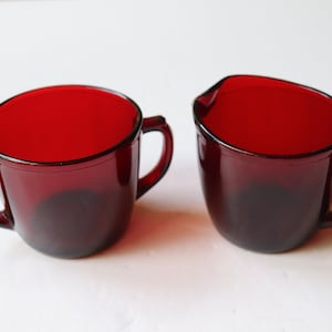 May include: Two ruby red glass vessels, one featuring a rounded cup shape with a side handle and the other a creamer pitcher with a pouring spout and a side handle. Both items are made of translucent, deep red glass with smooth, curved edges.