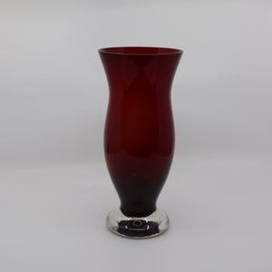 May include: A deep red glass vase with a smooth, curved silhouette that flares outward at the top. The vase features a solid, clear glass base that provides stability, creating a contrast between the vibrant crimson body and the transparent foot.