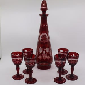 May include: A ruby red glass decanter with a stopper and six matching wine glasses. The set features etched white floral and geometric designs. The decanter is approximately 12 inches tall, and the glasses are about 4 inches tall.