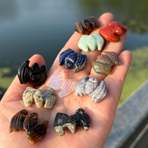 Könnte beinhalten: Eine Sammlung kleiner, aus Stein geschnitzter Fledermausfiguren in verschiedenen Farben und Materialien, darunter Schwarz, Blau, Grün, Rot, Braun und durchscheinendes Weiß, die in einer offenen Handfläche liegen.