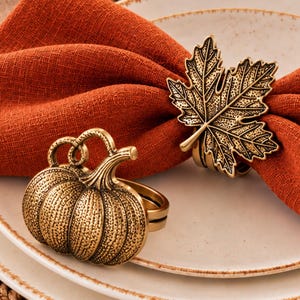 May include: A burnt orange fabric napkin folded and secured with a metallic gold-toned maple leaf napkin ring, accompanied by a separate metallic gold-toned pumpkin napkin ring resting on a stack of white ceramic plates with speckled edges.