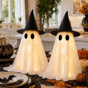 May include: Two illuminated ghost figures draped in white fabric, each wearing a pointed black witch hat with oval black eyes. These decorative pieces are positioned on a dining table surrounded by black and white pumpkins and autumn leaves.