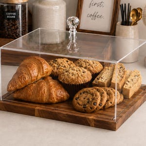 May include: A rectangular clear acrylic food display case with a crystal-style knob handle, set on a dark wooden base. The case contains two croissants, two muffins, and several chocolate chip cookies and biscotti arranged on the wooden tray.