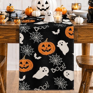 May include: A black table runner featuring a repeating pattern of orange jack-o'-lanterns, white ghosts, white spiderwebs, and the text "BOO!" in white. The runner is made of fabric and displays a festive Halloween theme with various seasonal motifs.