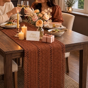 May include: A long, rectangular, rust-colored crochet table runner with an open-weave texture draped over a dark wooden dining table. The runner features vertical stripes of intricate, repeating patterns and hangs over the edge of the table.