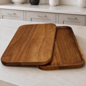 May include: Two rectangular wooden serving trays with rounded corners and a smooth, medium-brown wood grain finish. The trays are stacked slightly offset, showing a flat top surface on one and a recessed interior area on the other.